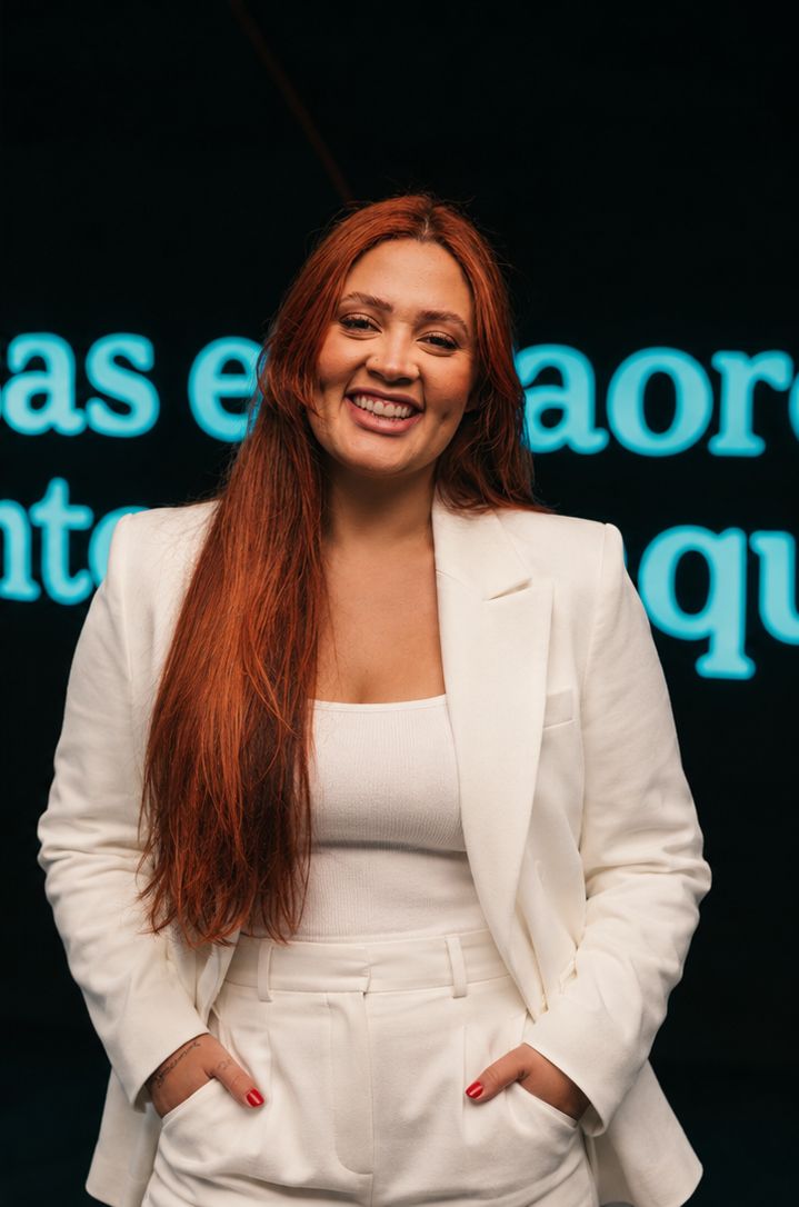 Natty Halls de pé, de terno branco, sorrindo, com as mãos nos bolsos, diante de um letreiro em neon